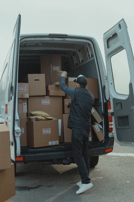A man wearing a grey cap, black gloves, a black quilted vest over a blue long-sleeved shirt, and black trousers stands outside a residential property next to a white moving van with a partially visibl