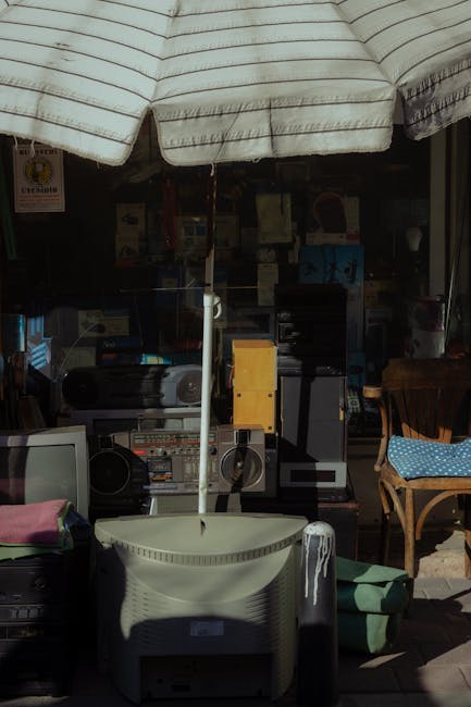 A collection of discarded electronic appliances and miscellaneous items arranged outdoors beneath a large, striped fabric awning. Visible objects include an old CRT television, a desktop computer monitor, a black stereo system with speakers, a yellow electrical box, and various computer peripherals. The items are placed on a concrete surface, with some stacked or leaning, suggesting they are ready for collection or disposal as part of a home relocation or furniture transport process. Behind the items, there is a background of shelves and storage units, indicating an indoor or semi-indoor space, possibly a shop or storage area. The scene is illuminated by natural daylight, casting shadows on the objects. Occasionally used in the context of removals, this image reflects the process of clearing, packing, and preparing bulky or electronic items for pickup or transportation, consistent with services offered by Man with Van Heston.