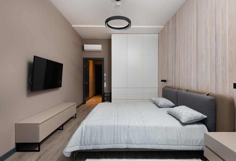 A modern bedroom interior with a double bed featuring a grey upholstered headboard and two light grey patterned pillows on a white bedsheet. To the left of the bed, there is a white, low-profile media unit with black legs, positioned against a beige wall. A flat-screen television is mounted on the wall above the media unit. At the foot of the bed, a white wardrobe with smooth, handleless doors is visible, with a small black stool placed beside it near a dark doorway leading into a hallway. An air conditioning unit is installed above the doorway on the beige wall. The room has a light wood laminate floor and a contemporary black circular ceiling light fixture. The environment appears well-lit with natural and artificial light, indicating a clean, tidy, and well-maintained space, suitable for house removals or packing and moving services in Heston as provided by Man with Van Heston.