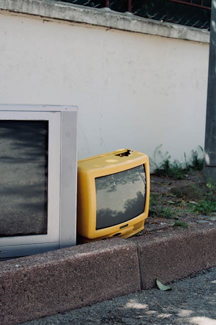 A collection of discarded electronic appliances and miscellaneous items arranged outdoors beneath a large, striped fabric awning. Visible objects include an old CRT television, a desktop computer monitor, a black stereo system with speakers, a yellow electrical box, and various computer peripherals. The items are placed on a concrete surface, with some stacked or leaning, suggesting they are ready for collection or disposal as part of a home relocation or furniture transport process. Behind the items, there is a background of shelves and storage units, indicating an indoor or semi-indoor space, possibly a shop or storage area. The scene is illuminated by natural daylight, casting shadows on the objects. Occasionally used in the context of removals, this image reflects the process of clearing, packing, and preparing bulky or electronic items for pickup or transportation, consistent with services offered by Man with Van Heston.