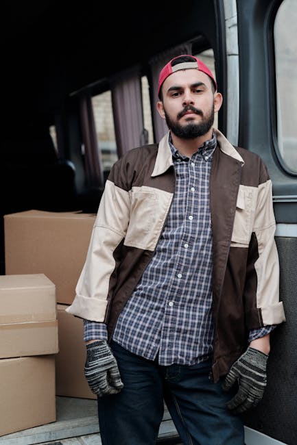 A man with a beard and wearing a red baseball cap, a brown and beige jacket over a checked shirt, and grey work gloves stands inside a van during a home relocation process. Behind him, the van's inter