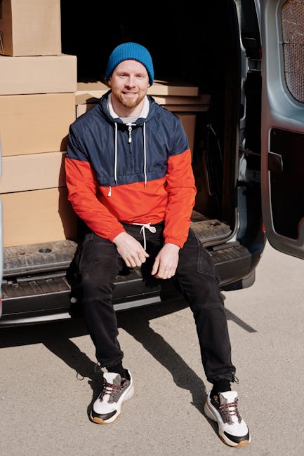 A man with a beard and wearing a red baseball cap, a brown and beige jacket over a checked shirt, and grey work gloves stands inside a van during a home relocation process. Behind him, the van's inter