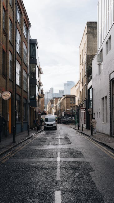 A narrow urban street scene with a wet, dark asphalt road marked with a central white line, flanked by modern multi-storey buildings with large windows, some featuring balconies and external air conditioning units. Several black bollards line the sidewalks on both sides of the street, with a white van parked near the center and a few pedestrians walking along the pavement. In the background, taller city buildings are visible under a cloudy sky, creating a typical cityscape environment suitable for home relocation and furniture transport. The scene is well-lit by natural daylight, and the image reflects a typical setting where a professional removals company such as Man with Van Heston might perform moving services, including packing, loading, and transport of household items along tight city streets.