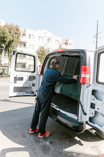 A person wearing black overalls and red shoes is loading or unloading a large green cardboard box into the rear cargo area of a white panel van with side door and rear doors open. The van is parked on a street near a sidewalk, with multi-storey residential buildings and trees visible in the background. The scene is set during daylight with clear weather, and the individual appears to be engaged in a home relocation activity, possibly part of furniture transport or packing and moving process managed by Man with Van Heston. The van's interior is empty except for the person's activity, and the open doors reveal space suitable for moving boxes or household items. The image captures a moment of the loading process in an urban environment, illustrating professional removals or home relocation services.
