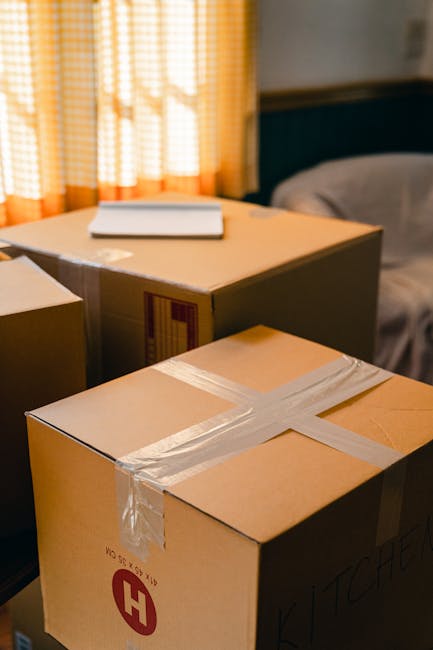 A delivery truck with a white cargo area is parked on a city street during daylight hours, loaded with cardboard boxes wrapped in plastic and secured with straps, positioned near an ornate multi-store