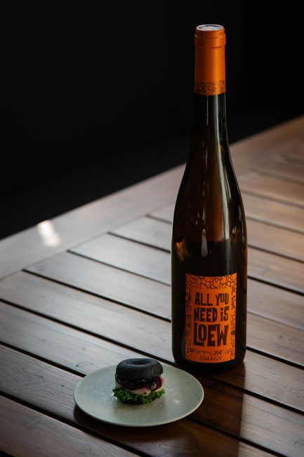 A tall, dark glass bottle of wine with an orange cap and label featuring the text 'All You Need Is LOEW' is positioned on a wooden table. Next to the bottle, there is a small white plate holding a canapé topped with black and pink ingredients, garnished with a small sprig of herbs. The table is indoors, with natural light casting soft shadows, and appears to be part of a home setting. This setup reflects elements involved in home relocation preparations, including packing and transporting beverages, as handled by companies like Man with Van Heston, who specialise in removals and moving services.