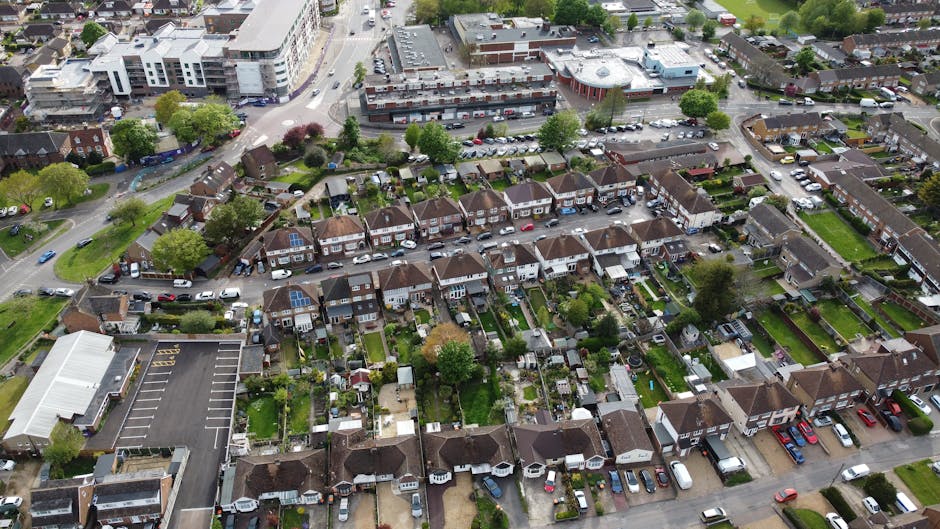 An aerial view of a residential neighbourhood showing a row of terraced houses with small gardens, some with sheds or patios, and several parked cars along the street. To the left, a large parking area with marked parking bays is visible adjacent to a commercial building. The scene also includes green spaces with trees and shrubs dispersed throughout. In the background, larger modern buildings and a busy street with moving vehicles can be seen. The overall setting depicts a typical urban area transitioning from residential to commercial zones, with a focus on the housing layout and vehicle access, supporting furniture transport, home relocation, and moving logistics activities, as handled by companies like Man with Van Heston.