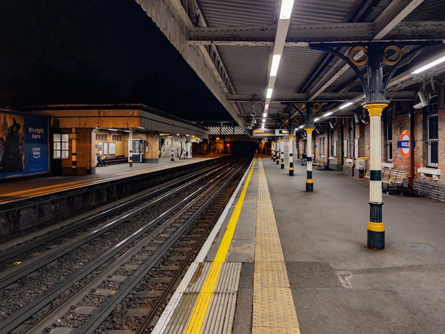 A nighttime scene at Hounslow West railway station platform showing empty train tracks, illuminated by overhead lighting. The platform features a tactile paving strip near the edge for safety, black and yellow striped bollards along the platform perimeter, and brick station walls with signage and benches. On the left side, a small shelter with a person sitting inside is visible, while on the right, there are brick walls, windows, and station signage. The environment appears quiet, with no passengers present, and the area is prepared for the loading process involved in house removals and furniture transport, such as potential loading of furniture or boxes onto transport vehicles, consistent with home relocation services provided by Man with Van Heston.
