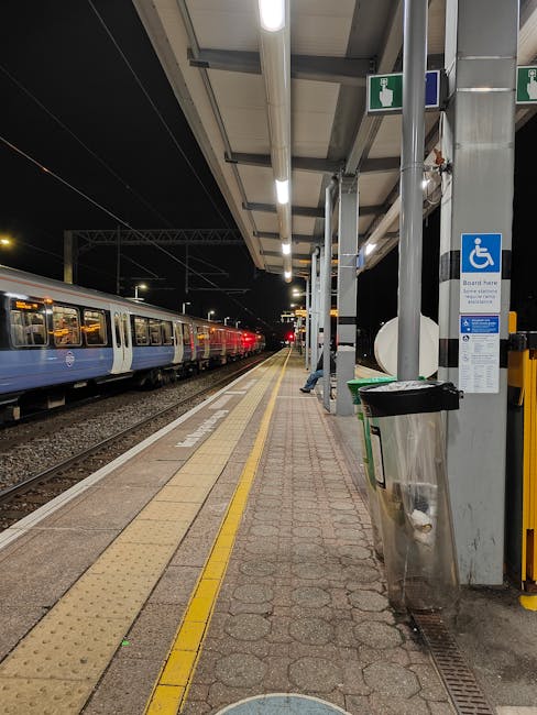 View of Hounslow West train station platform at night showing a stationary train with blue and white carriages on the left side, illuminated by overhead lighting. The platform features tactile paving strips and yellow warning lines along the edge. On the right side, there is a covered waiting area with metal supports, a blue accessible parking sign, and a metal waste bin. The station's surroundings are dark, highlighting the artificial lighting, and no people are visible in the image. This scene reflects the typical environment encountered during home relocation or furniture transport processes, illustrating the station's infrastructure as part of moving logistics, as managed by Man with Van Heston.