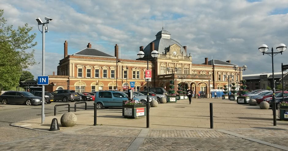 A nighttime scene at Hounslow West railway station platform showing empty train tracks, illuminated by overhead lighting. The platform features a tactile paving strip near the edge for safety, black and yellow striped bollards along the platform perimeter, and brick station walls with signage and benches. On the left side, a small shelter with a person sitting inside is visible, while on the right, there are brick walls, windows, and station signage. The environment appears quiet, with no passengers present, and the area is prepared for the loading process involved in house removals and furniture transport, such as potential loading of furniture or boxes onto transport vehicles, consistent with home relocation services provided by Man with Van Heston.