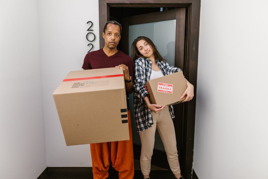 A man and a woman standing inside a doorway with a dark wood frame, both holding cardboard boxes for a home relocation. The man, dressed in a maroon shirt and orange trousers, is holding a large cardb
