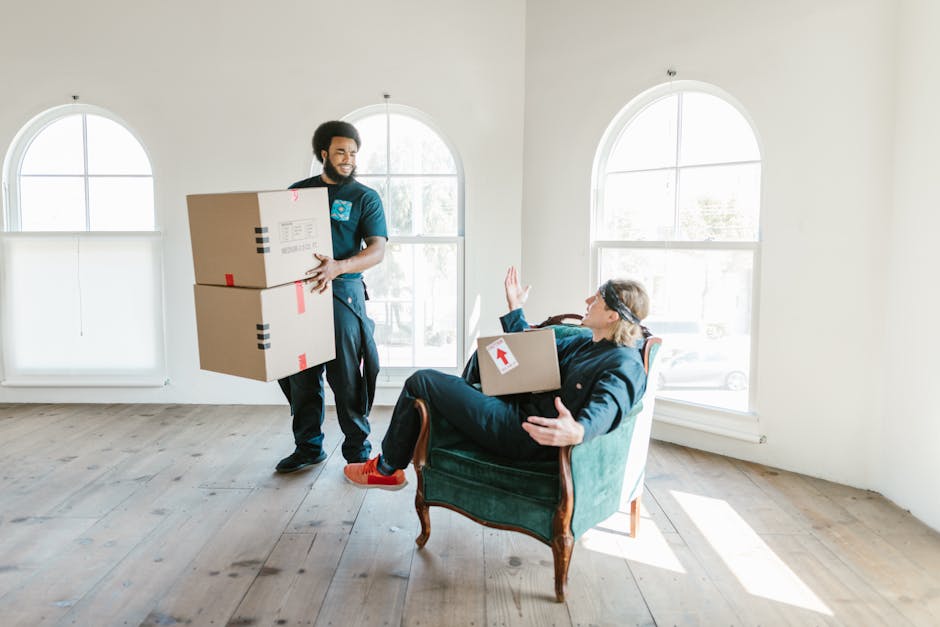 A man and a woman standing inside a doorway with a dark wood frame, both holding cardboard boxes for a home relocation. The man, dressed in a maroon shirt and orange trousers, is holding a large cardb