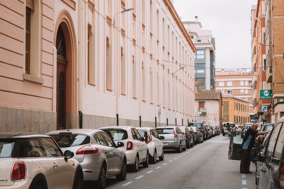 A narrow urban street scene in Heston with parked cars lining both sides of the road, including compact hatchbacks and larger vans, some with open doors. The street is bordered by a large, light-pink historic building with arched windows and decorative architectural details, and a mix of modern and older buildings in the background. A man is seen loading or unloading a vehicle on the right side, with packing materials and plastic wrap visible in the vicinity. The pavement and street markings are clear, indicating a restricted parking zone that may impact home relocation or furniture transport activities. The scene captures the typical challenges of moving logistics in tight city streets, relevant to professional removals provided by Man with Van Heston, inclusive of parking restrictions and urban navigation considerations in Heston.