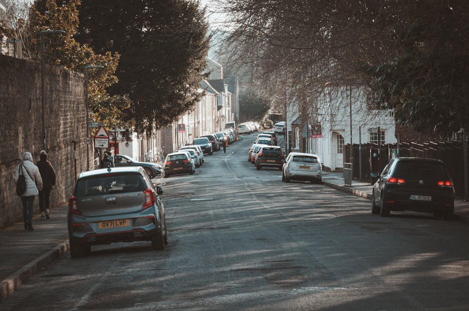 A residential street scene in Heston during daytime, featuring parked cars lining both sides of the narrow road, with some vehicles parked partially on the pavement and others along the curb. The street has a slight incline, and the road surface shows patches and wear. On the left, two pedestrians are walking past, carrying bags; they are dressed in coats suitable for cool weather. To the right, a black van is parked, and a white house with a white door and window is visible behind it. In the background, more parked cars and trees with sparse foliage can be seen, indicating late autumn or early winter. The scene appears well-lit with natural daylight, with some shadows cast by trees and vehicles. This setting illustrates urban house relocation logistics, where careful vehicle and furniture transport planning is necessary due to parking restrictions and narrow streets, as addressed by Man with Van Heston in their removals services.