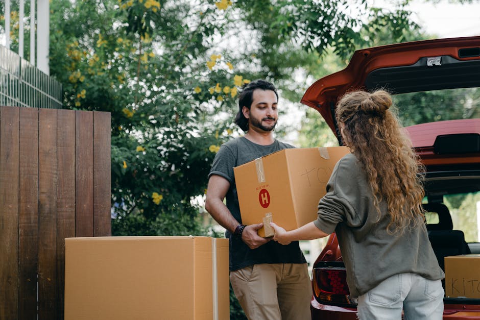 A man with dark hair and a beard, dressed in a grey t-shirt and beige trousers, is standing outdoors next to a red vehicle with its trunk open. He is receiving a medium-sized cardboard box from a woman with long, curly hair, who is wearing a grey hoodie and light-colored pants. The woman is handing over the box directly to the man, and additional cardboard boxes are visible on the ground nearby, some sealed with packing tape. The scene takes place in a garden or yard area, with a wooden fence on the left and lush green trees and foliage in the background, indicating an outdoor home relocation or packing process supported by Man with Van Heston. The lighting is natural, suggesting daytime, and the overall setting reflects typical furniture transport and packing activities involved in house moves.