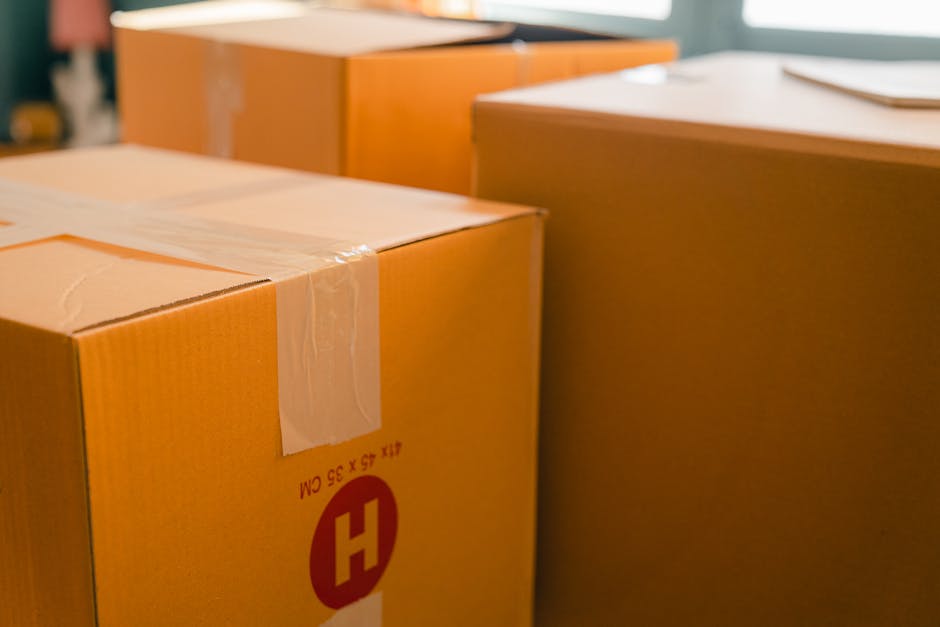 Close-up view of several cardboard moving boxes, sealed with clear packing tape, arranged inside a room, with some placed on a flat surface and others on the floor. The boxes are partially visible, showing the edges and corners, with a focus on one box in the foreground featuring a red 'H' logo and size markings. The background includes a glimpse of furniture such as a wooden cabinet and a desk, indicating a home environment prepared for a house removal. The lighting is natural, and the scene captures the packing stage of a furniture transport process, typical of home relocation services provided by Man with Van Heston, supporting efficient packing and loading during household moves.