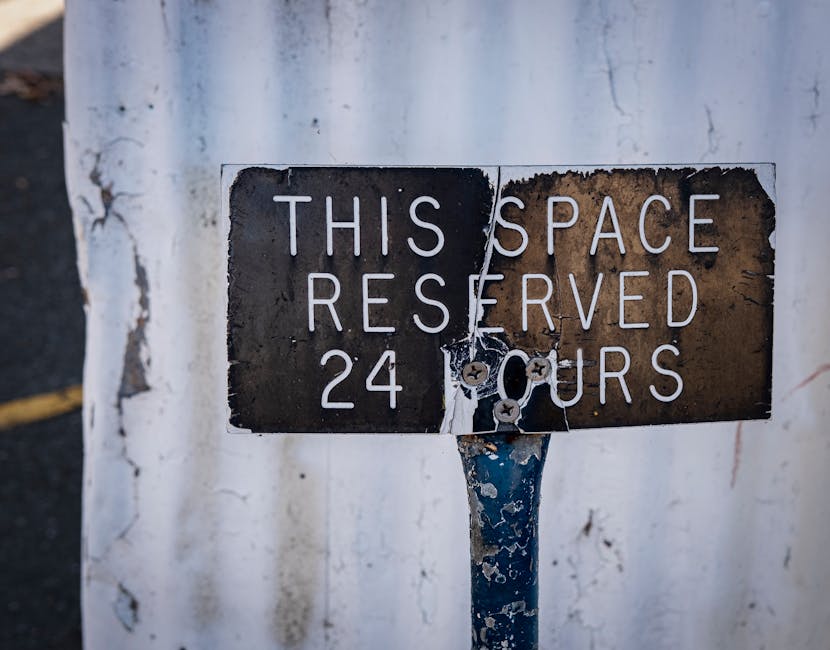 Close-up image of a weathered metal sign attached to a metal post, with peeling paint and rust. The sign reads 'THIS SPACE RESERVED 24 HOURS' in white capital letters on a black and brown background, indicating restricted parking or entry. The background features a white wall with visible peeling paint and minor cracks, typical of an exterior urban environment. This setting reflects conditions often encountered during house removals or moving logistics, where parking restrictions and waste rules can impact a home relocation process. Man with Van Heston provides removals services including loading and furniture transport, often requiring careful adherence to local regulations such as those indicated by parking restriction signs.