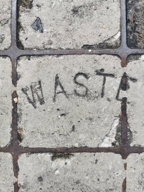 Close-up view of a paved outdoor area showing a stone brick with the word 'WASTE' engraved in it, surrounded by other irregularly shaped stones and concrete paving—part of a walkway or yard. The stones have a weathered appearance with some chips and dirt visible on their surfaces. The engraved brick is positioned near the center of the image, indicating the area is designated for waste disposal within a property. This setting is relevant to house removals and moving services provided by Man with Van Heston, as it highlights considerations for waste disposal and logistics during home relocation or furniture transport. The lighting is natural, suggesting an outdoor environment, with a focus on the textured surface of the paving stones and the engraved brick that marks waste management areas as part of a broader moving or clearance process.